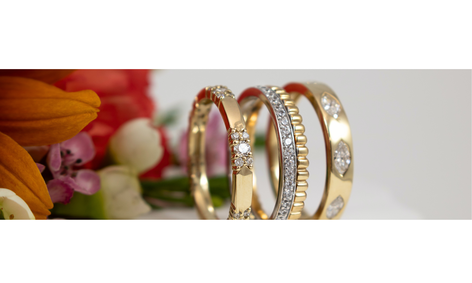 Three gold and silver rings with clear gemstones, displayed in a close-up shot with blurred orange and pink flowers on the left side.